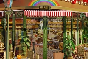 빨간색과 흰색 줄무늬 차양 아래 무지개 장식이 있는 베이커리 전경, 식물들과 테이블이 배치되어 있다