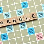 가로로 놓인 나무 블록에 SCRABBLE이라는 글자가 써 있고, 점수가 표시된 바둑판 무늬의 게임 보드 위에 놓여있다